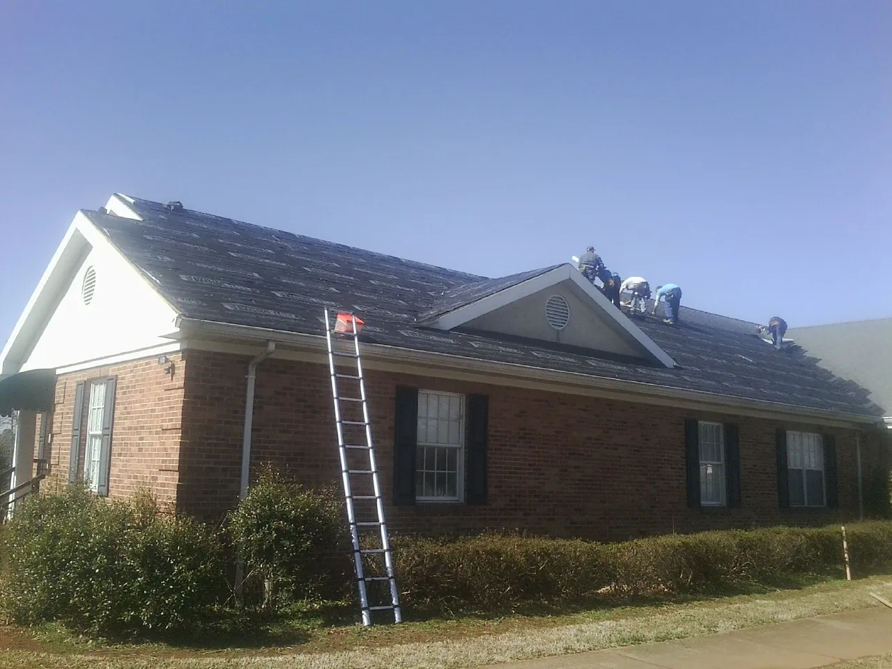 Crew tearing shingles off a brick ranch roof with a ladder set — Metal Roof Repair work by our crew in Milford